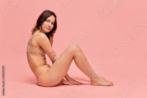 Portrait of young, beautiful, slim girl in beige underwear posing against pink studio background. Self-care and acceptance. Concept of body and skin care, figure, fitness, health, wellness.