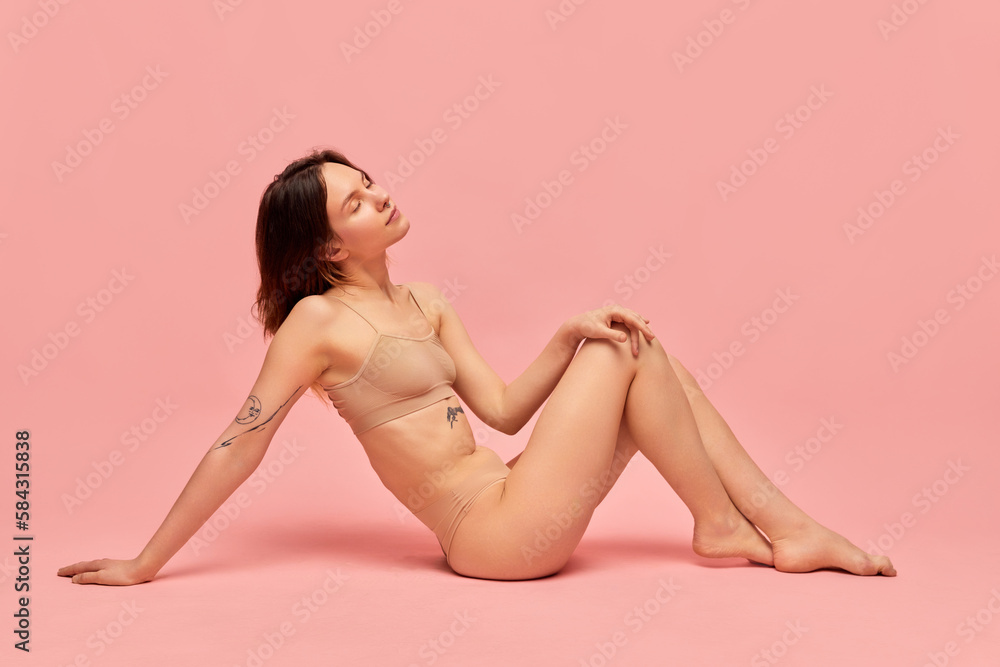 Fototapeta premium Portrait of young relaxed girl with slim healthy body in beige underwear posing against pink studio background. Natural beauty. Concept of body and skin care, figure, fitness, spa, health, wellness.