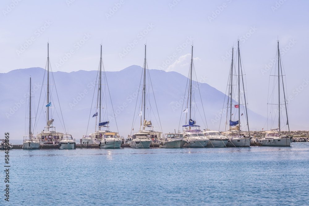 Several yachts moored in one row in a light morning haze against the backdrop of a mountain in the port of Budva, Montenegro. Gentle seascape with single-masted passenger boats in the Adriatic Sea