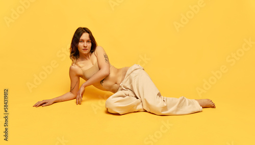 Attractive young girl with skinny body posing in top and jeans against yellow studio background. Tattoo and piercing. Concept of beauty, body and skincare, figure, fitness, health, wellness.