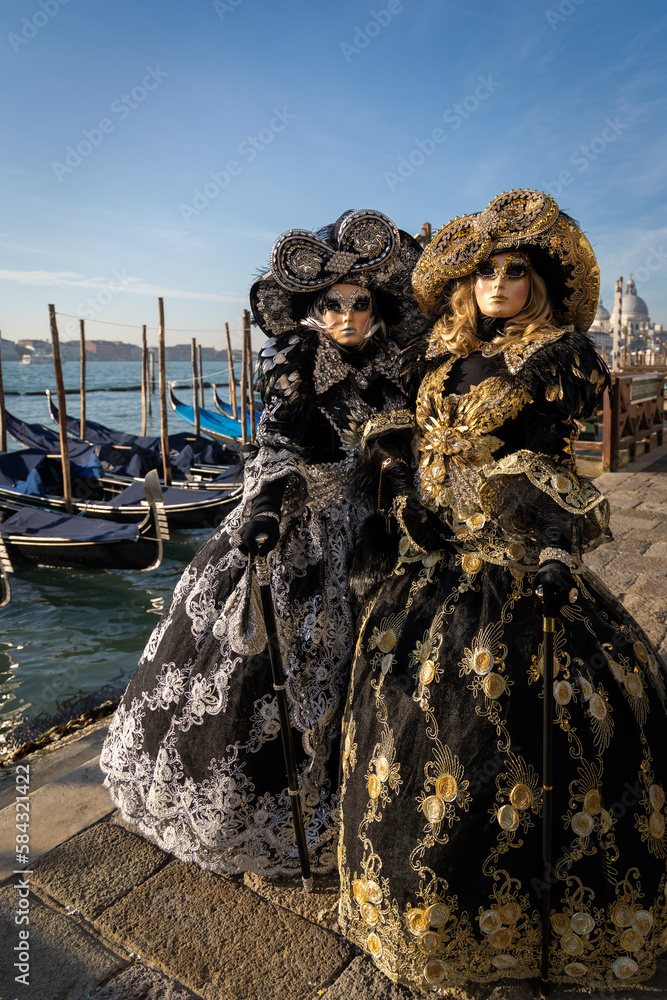Venice carnival costumes 2023 , unrecognizable two masked models posed near a canal in Venice. Vintage costumes with blue skys and sea and gondala's to the background .