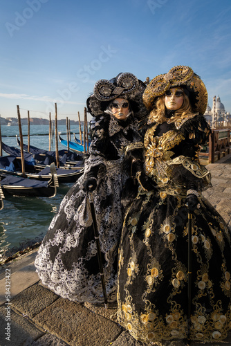 Venice carnival costumes 2023 , unrecognizable two masked models posed near a canal in Venice. Vintage costumes with blue skys and sea and gondala's to the background .