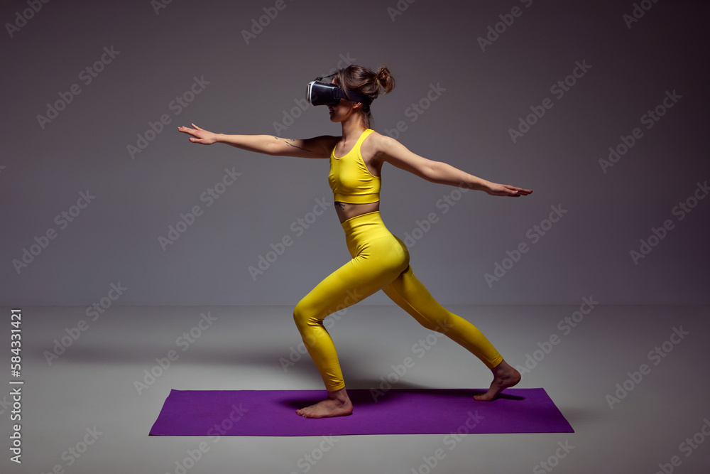 Fototapeta premium Young slim girl in yellow sportswear training, doing yoga exercises in VR glasses against purple background. Flexibility and strength. Concept of body , figure, fitness, health, wellness.