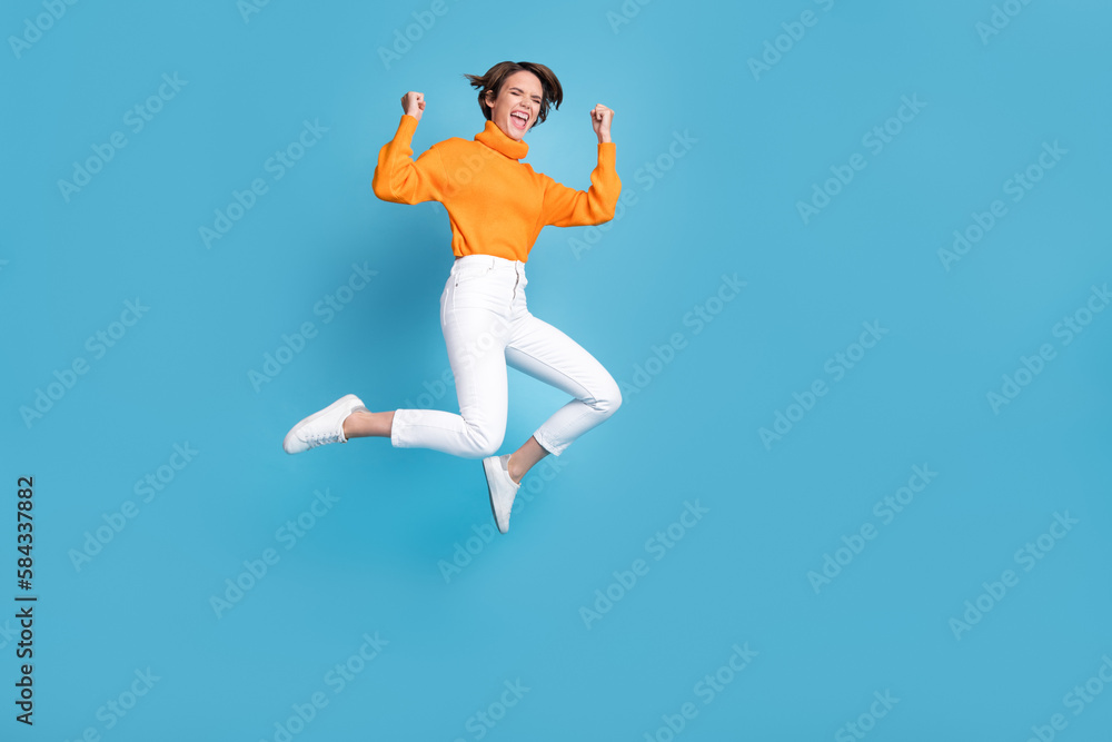 Full body photo of attractive young woman raise fists shout yeah jump crazy dressed stylish orange look isolated on blue color background