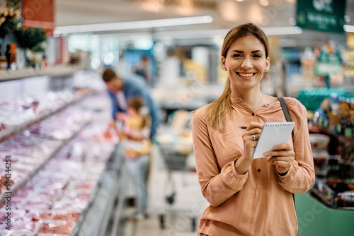 Slika na platnu Happy woman going through grocery list while shopping in supermarket and looking at camera