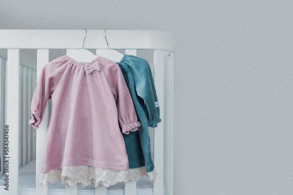 Pink, blue dresses for baby girl on white hanger near white bed in nursery. Set of baby clothes and accessories for birthday party. Fashion childs outfit. Copy space Pink, blue dresses for baby girl on white hanger near white bed in nursery. Set of baby clothes and accessories for birthday party. Fashion childs outfit. Copy space