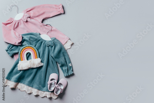 Blue, pink dress for baby girl with white hanger, rainbow toy, shoes on grey background. Set of baby clothes and accessories for birthday party. Fashion childs outfit. Flat lay, top view. Copy space