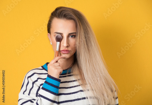 Happy young woman holding make-up brush, isolated on yellow background. Pretty girl