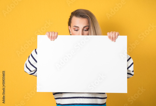 Close-up woman behind white board and looking down to empty space, isolated on yellow background. Pretty girl showing blank space for text near her head