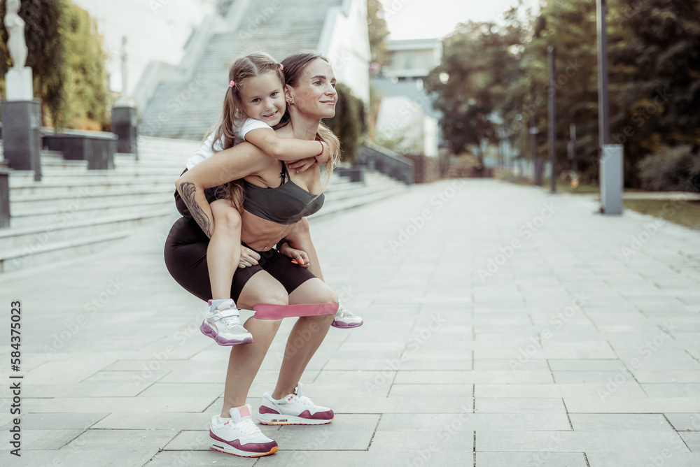 Obraz premium Athletic mom exercising with her little daughter on her back outdoors. Squat with fitness rubber bands. Healthy lifestyle, fitness active family concept. Training together