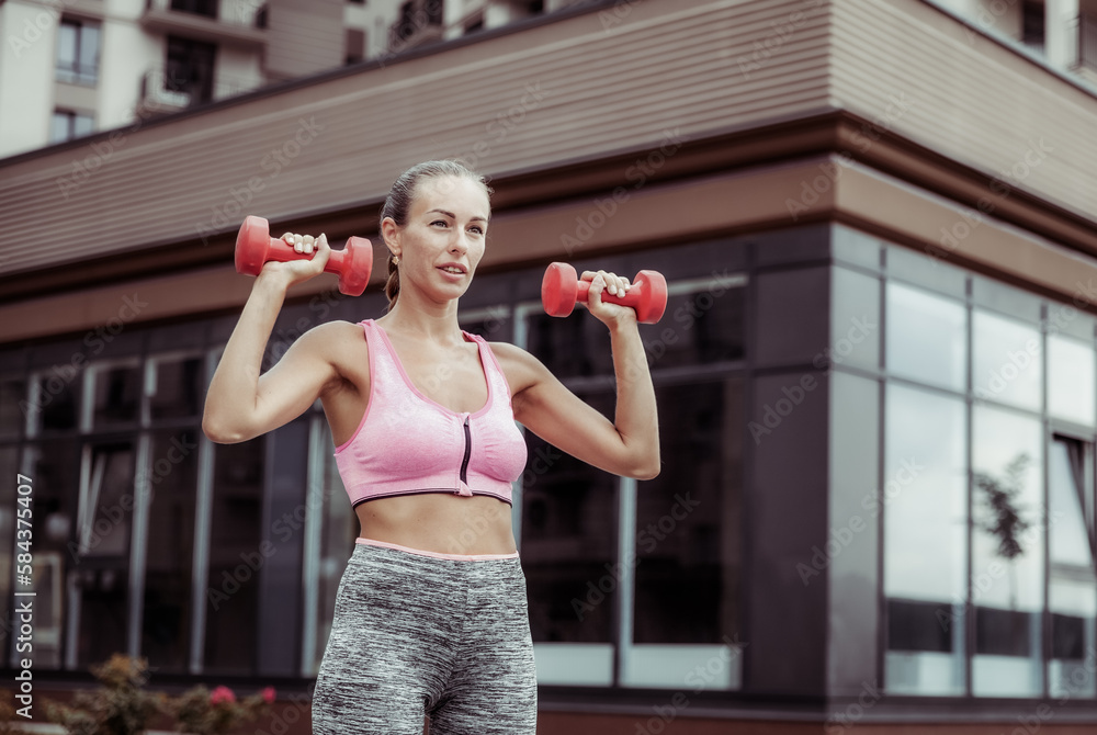Fototapeta premium Active lifestyle concept. Fit woman in sportswear trains muscles with dumbbells in hands outdoors