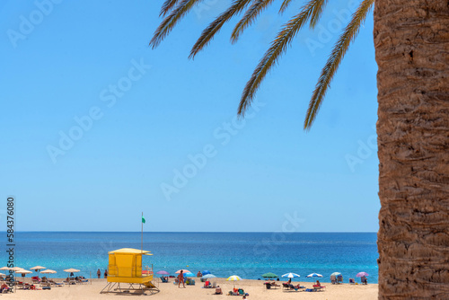 playa de Morro Jable de arena blanca y mar turquesa llena de gente disfrutando del sol junto a un puesto de socorrista amarillo cerca de una palmera y con bandera verde en Fuerteventura  Islas canaria