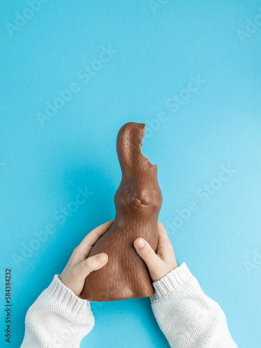 Chocolate Easter bunny with ears bitten off in hands of child on blue background, family concept. Vertical, top view, copy space