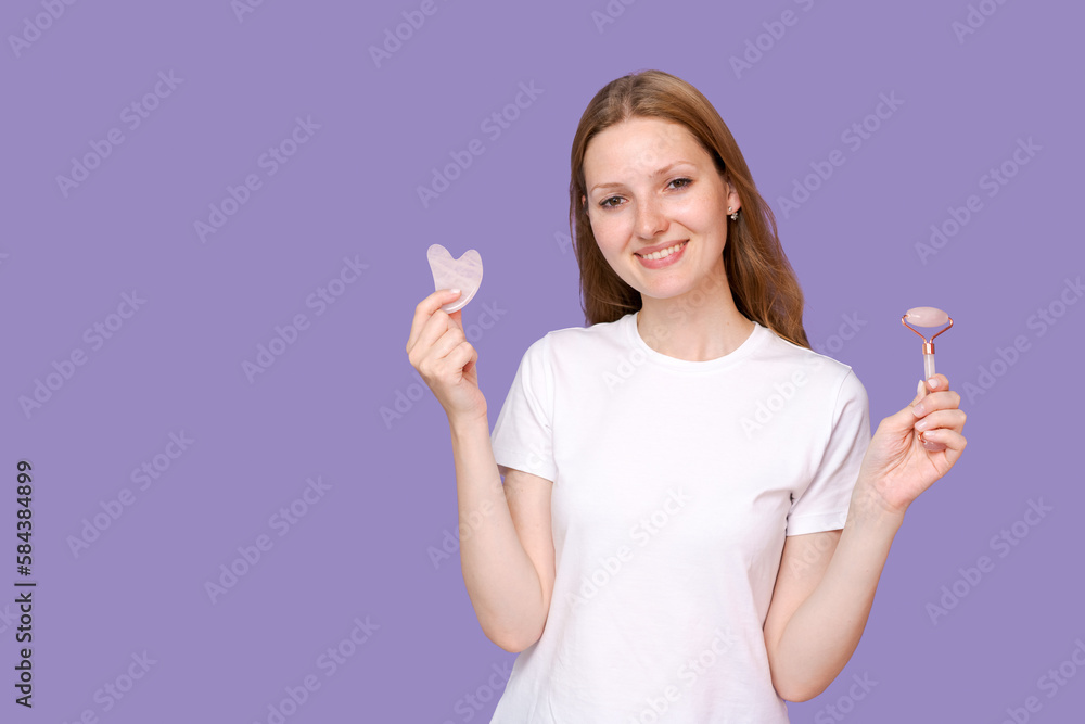 Young lady massaging her face by Gua Sha Quartz Stone Scraper for skin care, posing on purple background, wearing white t-shirt. A woman uses a natural massager. Facial massage, female beauty trends. Young lady massaging her face by Gua Sha Quartz Stone Scraper for skin care, posing on purple background, wearing white t-shirt. A woman uses a natural massager. Facial massage, female beauty trends.