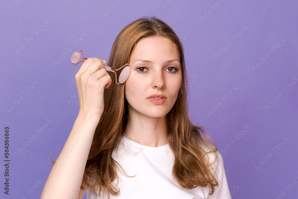 woman facial skin massage with natural guasha roller. Girl enjoy using lifting tool on her face, massaging muscles to reduce tension, rubbing skin care treatment in white t-shirt on purple background woman facial skin massage with natural guasha roller. Girl enjoy using lifting tool on her face, massaging muscles to reduce tension, rubbing skin care treatment in white t-shirt on purple background