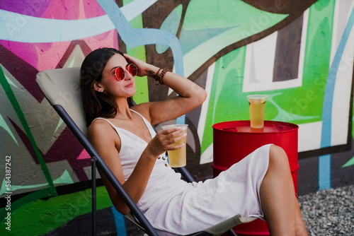 Three stylish young women dancing and laughing with drinks in hand against a colorful graffiti wall. A lively urban summer vibe full of joy, friendship, street art, and carefree energy.