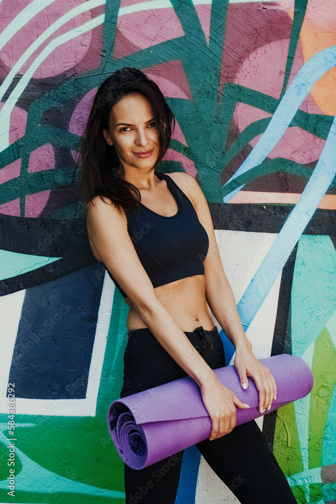 Summer portrait of pretty woman posing alone at city park after her yoga fitness class, sportive outfit, holding mat for exercises