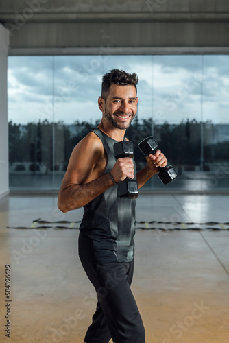 Portrait of a smiling caucasian male personal instructor, looking at camera, exercising with dumbbells in a gym. Workout lifting dumbbell weights. Vertical, window background.