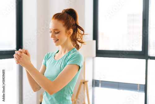 Side view of happy sporty young woman doing stretching exercise wrist before fitness training at home office near window. Positive redhead fit female warming up before muscles in apartment. Free Download Side view of happy sporty young woman doing stretching exercise wrist before fitness training at home office near window. Positive redhead fit female warming up before muscles in apartment. - GFXLoad