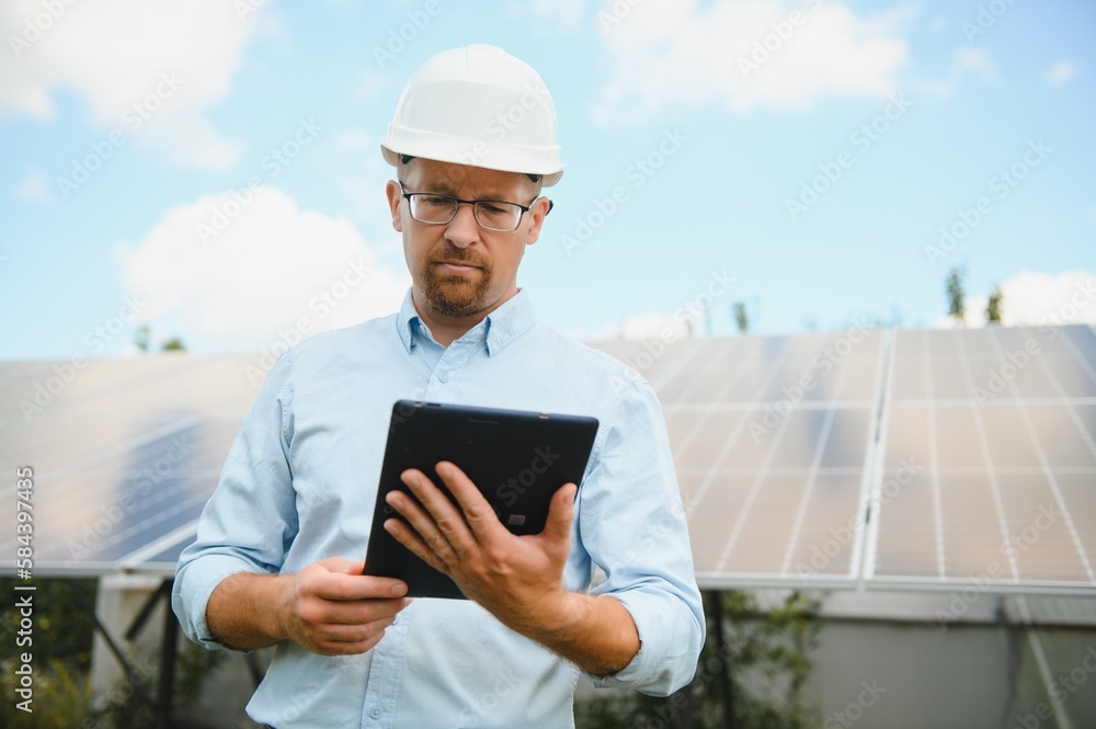 Portrait of male engineer with tablet in his hands near the solar panels station, wearing helmet at sunny day. Green ecological power energy generation.