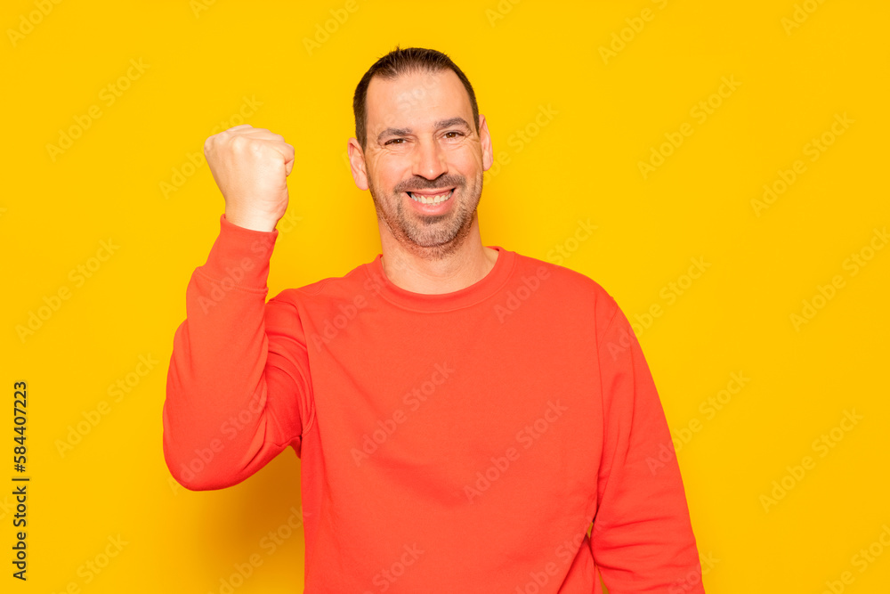 Bearded hispanic man in his 40s wearing a red jumper raising his fist and smiling in victory, isolated over yellow background. Bearded hispanic man in his 40s wearing a red jumper raising his fist and smiling in victory, isolated over yellow background.