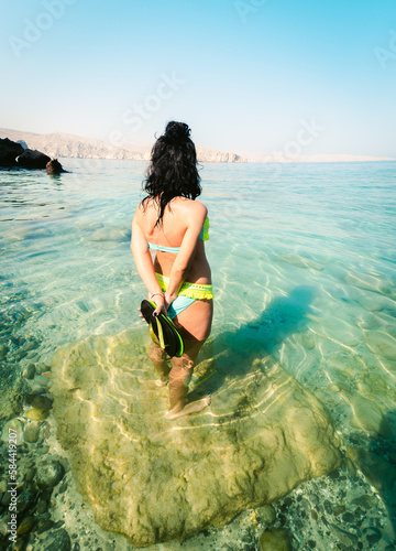Back view pretty tourist woman stand in clear turquoise water in persian gulf Mirellas island. Oman coastline paradise. Musandam.