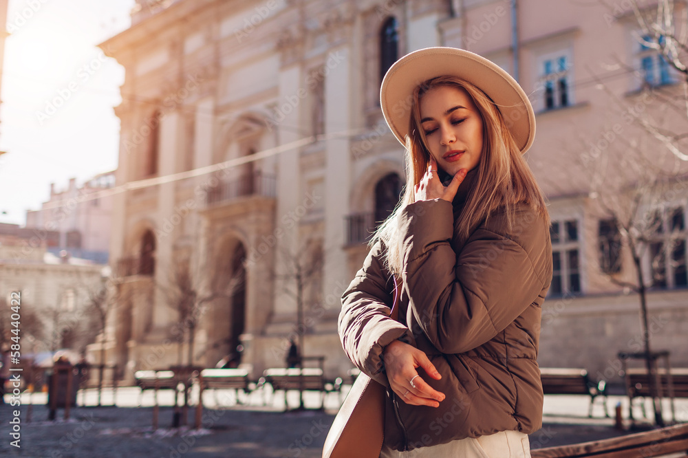Fototapeta premium Fashionable young woman walking on street of European city wearing stylish hat and jacket. Spring fashion