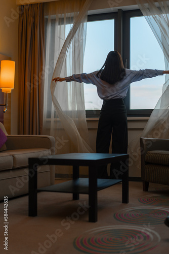 young woman opening curtain while standing in luxury hotel room looking through window enjoying city skyscrapers close up