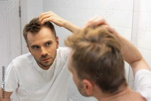 Middle aged caucasian white man with a short beard looks at his hair in the mirror in the bathroom and worried about balding. The concept of the problem of male hair loss, early baldness and alopecia photo