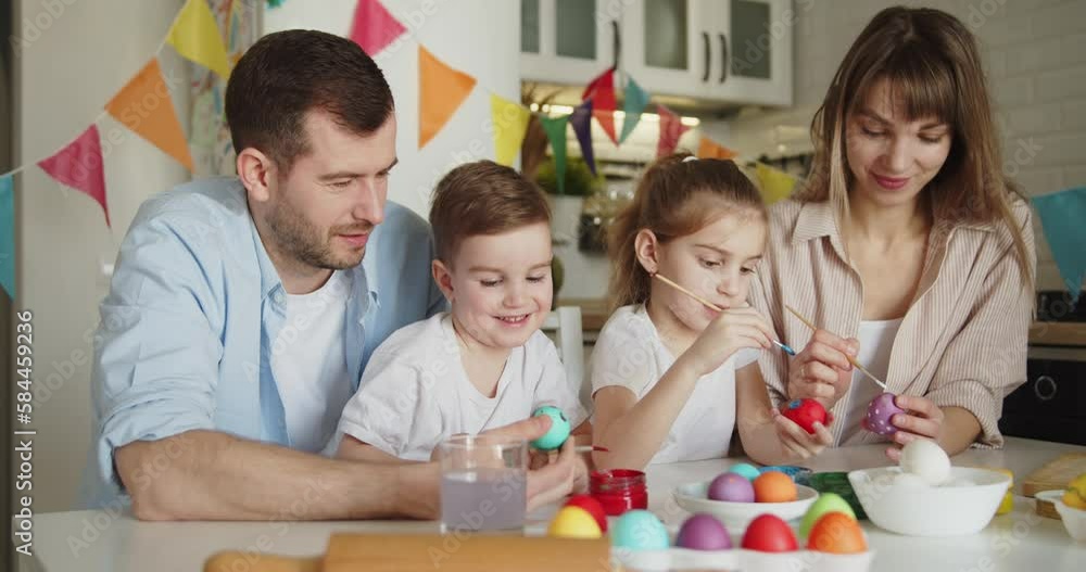 Joyful family of four painting Easter eggs together at the table in bright modern kitchen. Family gathering in preparation for Easter. Joyful family of four painting Easter eggs together at the table in bright modern kitchen. Family gathering in preparation for Easter.