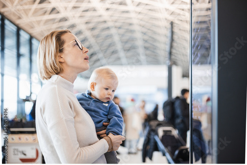 Mother traveling with child, holding his infant baby boy at airport terminal, checking flight schedule, waiting to board a plane. Travel with kids concept