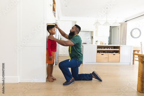 African american father and son measuring height against wall in kitchen photo