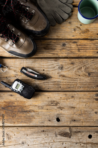 Camping equipment of trekking shoes, metal cup, walkie talkie on wooden background with copy space