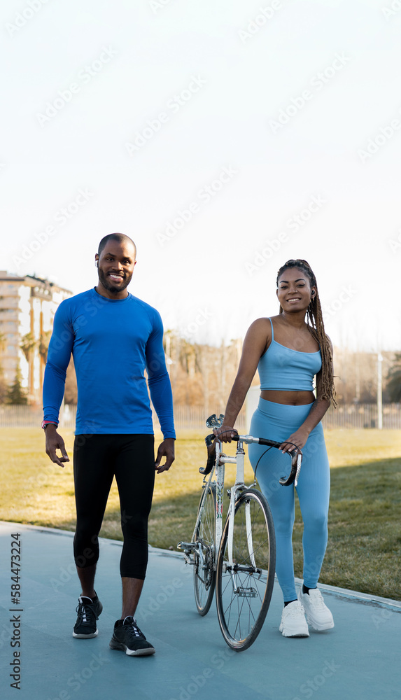 A dark-skinned couple is happily doing outdoor sports, the black man and the black woman with braids ride a bicycle in a park while looking at the camera. Concept of sport in couples.