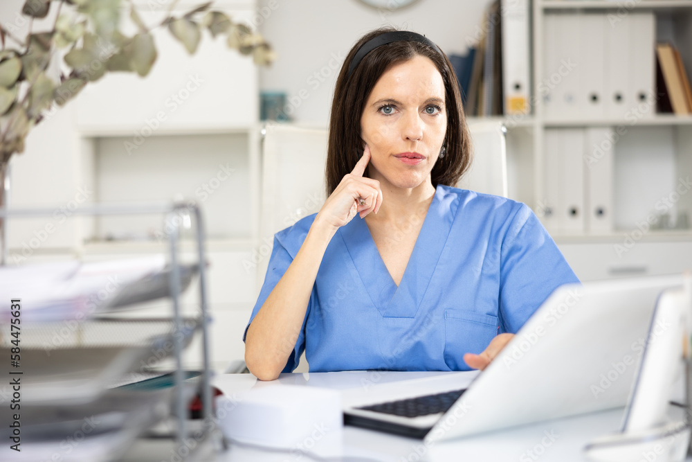 Portrait of female doctor in surgical scrubs sitting at working table in her office.