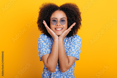 Beautiful positive fashionable african american curly haired woman with blue glasses, wearing blue summer dress, posing at camera and smiling friendly while standing over isolated orange background © Kateryna