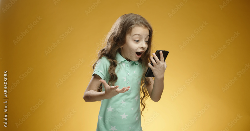 Funny Caucasian child holding and looking at phone, expressing an emotion of joy, surprised and shocked with open mouth isolated on a yellow background close-up. Copy space. Funny Caucasian child holding and looking at phone, expressing an emotion of joy, surprised and shocked with open mouth isolated on a yellow background close-up. Copy space.
