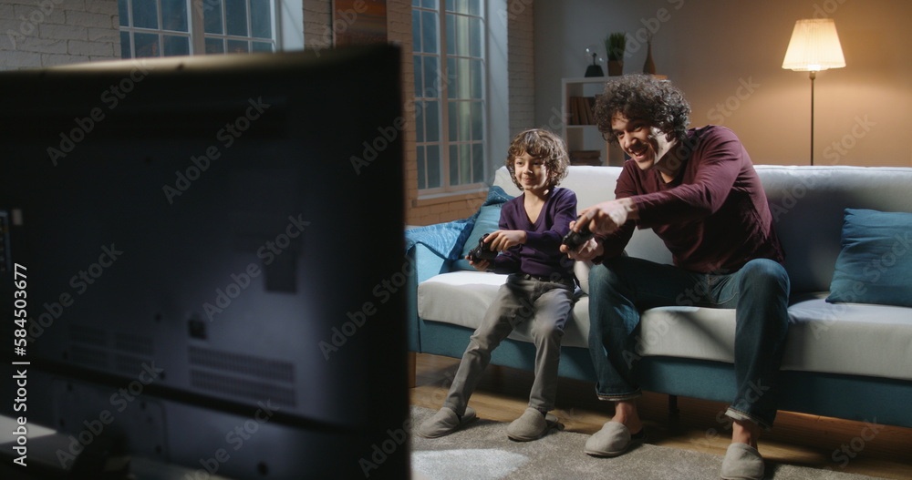 cheerful father and son playing video game at home. Father And Son Sitting On Sofa In Lounge Playing Video Game. Happy family concept. cheerful father and son playing video game at home. Father And Son Sitting On Sofa In Lounge Playing Video Game. Happy family concept.