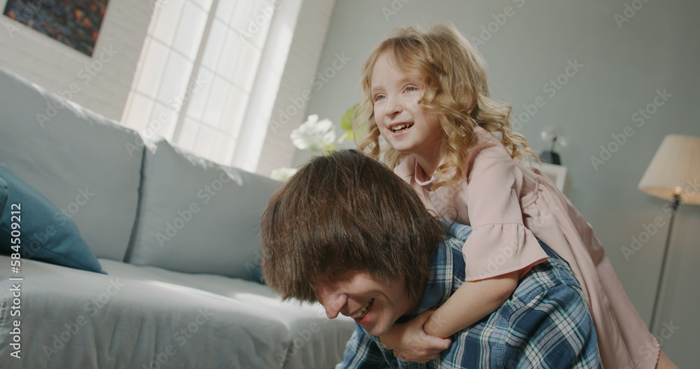 Funny joyful little caucasian girl having a piggy back ride on her father while he is doing push ups, spending time at home together - real people, family home Funny joyful little caucasian girl having a piggy back ride on her father while he is doing push ups, spending time at home together - real people, family home