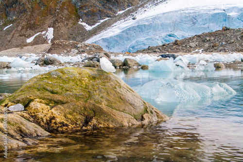 Semi circular ice ball balancing on a soft green mossy rock in the Arctic with a light blue glacier behind and Rocky Mountain behind that.