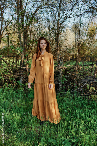 vertical photo of a beautiful, elegant woman in a stylish orange dress standing in the countryside in the shade of trees