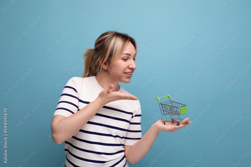 cute blond young mother showing shopping cart miniature on blue background with copy space cute blond young mother showing shopping cart miniature on blue background with copy space