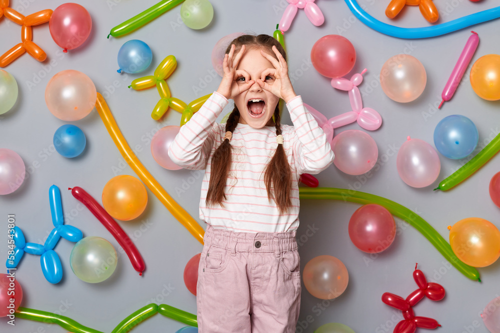 Fototapeta premium Portrait of excited little girl with braids wearing casual clothing making binocular gesture with hands, waiting for guests, standing against gray wall with colorful balloons.