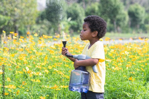 African 3 years old boy holding a big and heavy watering can, watering flowers garden on sunny day. Cute little child gardening, spraying water from a watering can to himself in summer. Funny moment photo