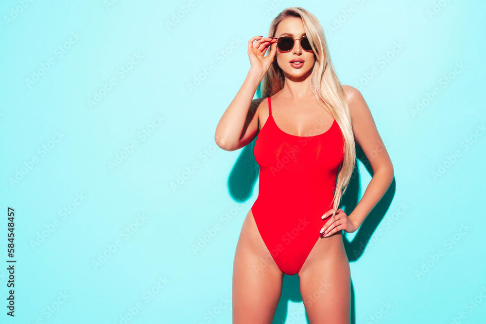Young beautiful sexy woman. Carefree model wearing pure red lingerie with big breasts. Hot tanned blonde posing near blue wall in studio in summer swimwear bathing suit. In sunglasses