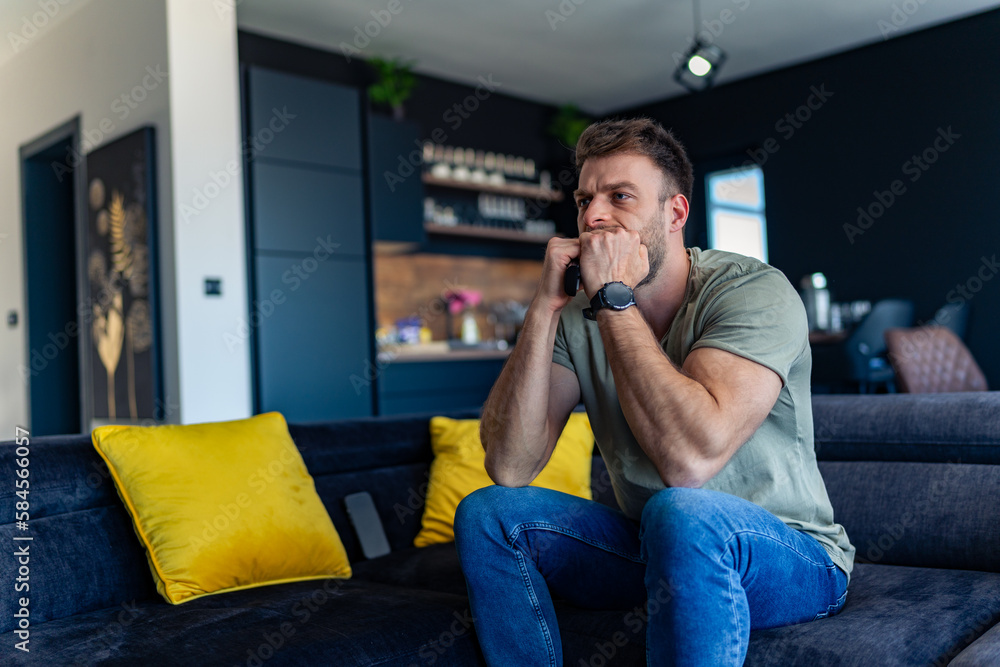 the young man is tense while watching the game, on the edge of the couch holding the remote control in his hand and getting annoyed the young man is tense while watching the game, on the edge of the couch holding the remote control in his hand and getting annoyed