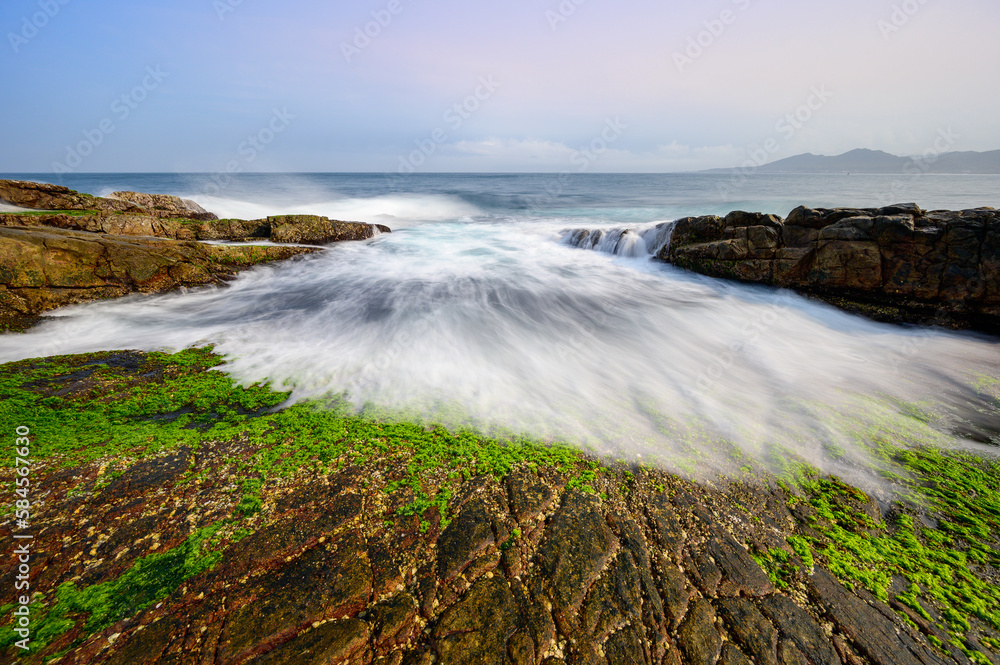 Fototapeta premium White waves wash over mossy brown rocks. Explore breathtaking coastal landscape. Clear Waters, and Stunning Views of the Pacific Ocean in New Taipei City.