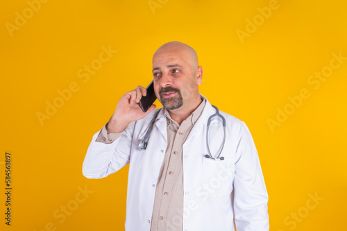 Telemedicine, close up portrait of middle aged bald doctor making telemedicine. Physician using smartphone. Isolated yellow background, copy space. Wearing white hospital coat, stethoscope.