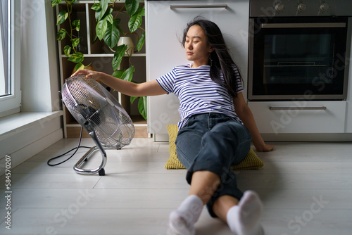 Overheating in homes. Tired overheated young Asian woman sitting on floor in kitchen near electric fan, cooling down at home during extreme summer heat, staying cool without air conditioning photo