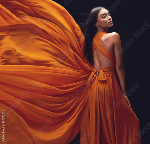 Fashion, woman and elegant, dress flying and style, beauty on dark background, portrait and model in orange in studio. Indian female, glamour and stylish with sexy person, luxury and designer wear